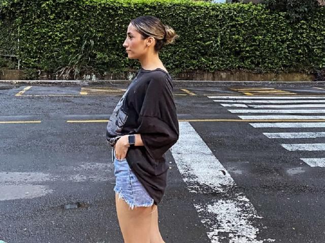AdharaCooperr, debout de profil, porte une chemise noire, un short en jean bleu, une montre noire et a les cheveux bruns attachés en chignon. Elle se trouve au niveau d'un passage piéton et regarde à gauche..