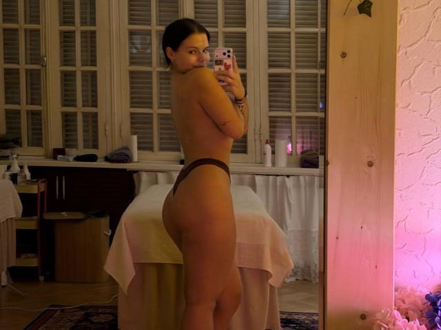 ClarenceGarin, topless, with brown thong, standing sideways, showing ass, taking mirror selfie, holding phone, in dimly lit room, wearing bracelet, with white windows and wooden wall.