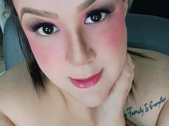 GiiannaMorrys has pink eyeshadow, glossy pink lips, a nose ring, and a tattoo on her shoulder. She's smiling with her hand on her chin, looking cute.