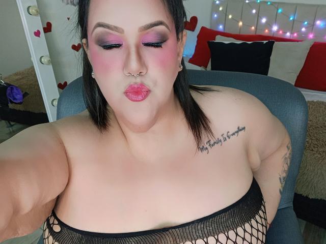 GiiannaMorrys with puffy pink lipstick, black fishnet top, nose ring, and tattoo on right shoulder, pouting, showing large tits, dark hair, and pink eyeshadow.