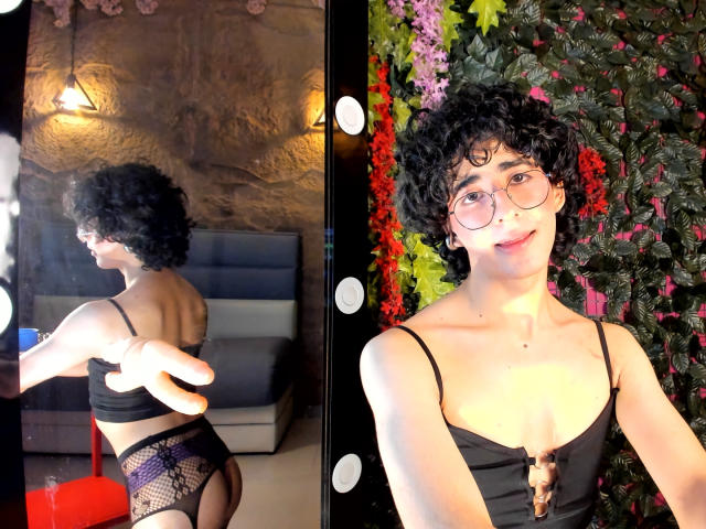ElliotNoir with curly black hair, glasses, and a black crop top, showing a bare butt in fishnet underwear, smiling at a mirror with colorful plant decor.