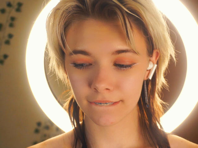 DestinyCassella, con i capelli biondi corti, l'eyeliner arancione e i denti argentati, indossa delle clip per le orecchie bianche e si trova di fronte a una luce circolare molto luminosa..