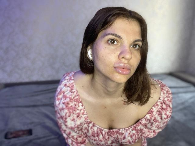 CicelyDence with pouty lips, wearing a pink floral top, white earbuds, brown hair, and light skin, looking up with a slightly curious expression. Her boobs are visible.