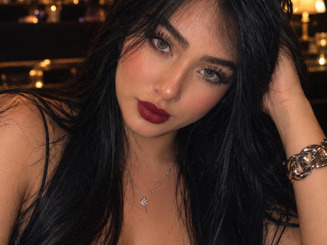 LilyConorr ha lunghi capelli neri, rossetto rosso e una collana d'argento con un fiore. Sta guardando la fotocamera con gli occhi verdi, e indossa un braccialetto vistoso al polso destro..