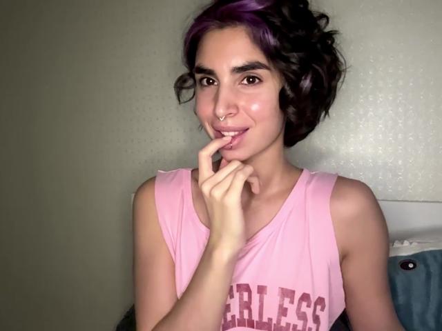 TameikaMacclellan with purple and black curly hair, nose ring, pink tank top, and finger in mouth, showing cute, shy expression. Soft light, subtle smile, pink lips.