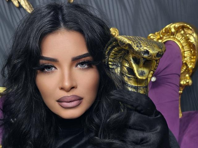 Devasha, con cabello negro largo, lápiz labial morado y pestañas postizas, mirando directamente a la cámara, vestida con una prenda negra, con una silla ornamentada en dorado y morado detrás de ella..