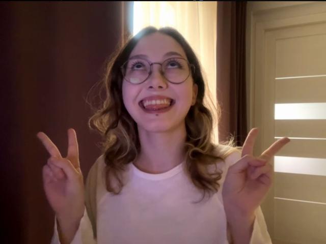 Rosario Vandagriff met golvend bruin haar, die een bril draagt en een witte blouse, lachend met haar tong uit, "V" tekens met haar handen, een klein neuspiercing, zachte binnenverlichting, casual, schattig, zelfverzekerd..