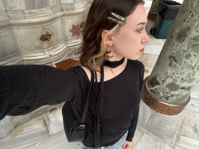 Rosario Vandagriff, avec ses cheveux blonds et bruns, son collier noir, sa chemise noire à manches longues et son sac bandoulière noir, prend un selfie, montrant son profil et ses boucles d'oreilles, avec une colonne en marbre..