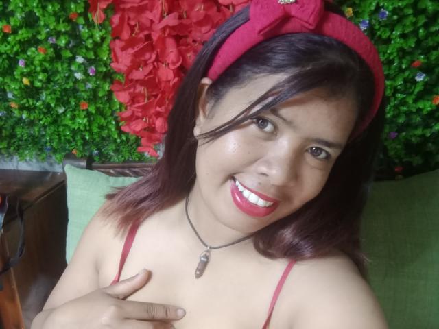 MariahSquirting69, cute Latina woman with red lipstick and dark hair, smiling, wearing red headband, necklace, and red bra, touching her small tits with right hand.