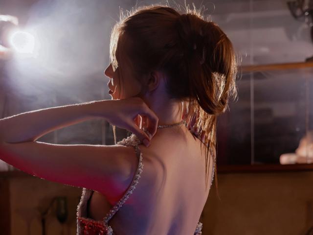 MargoKoli with her back to the camera, adjusting a sparkly dress, exposing her bare shoulders and upper back, in a dimly lit room with a bright light source.