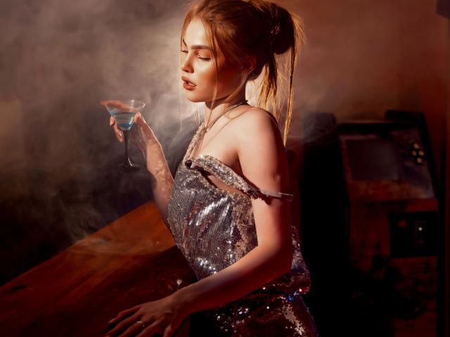MargoKoli in a shiny silver sequin dress, holding a blue cocktail, with smoke around her, her blonde ponytail, eyes closed, and hand on a wooden bar.