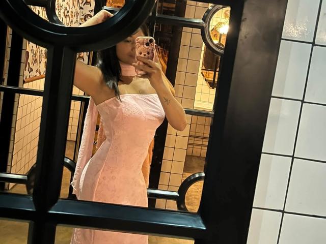 AshlyLee with her tits covered in a pink, strapless, textured dress, taking a mirror selfie with a black ring in her hair, holding a pink phone, in a tiled room.