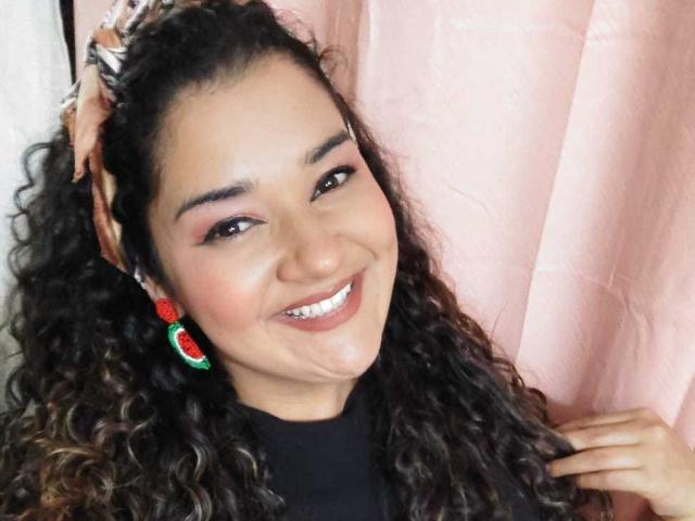 NataliaWendys with curly black hair, smiling, brown eyes, wearing red and green earrings, holding her hair up, black turtleneck, pink curtain background, showing her white teeth.