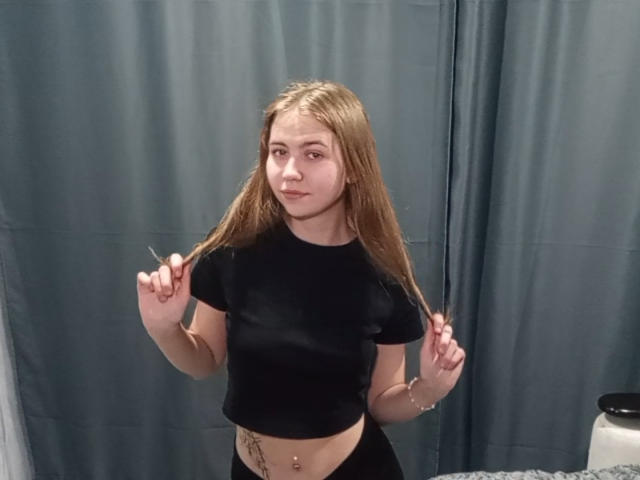 MargretWarriner with long blonde hair, black crop top, and black pants, holding her hair, showing a belly button piercing and a tattoo on her right side.
