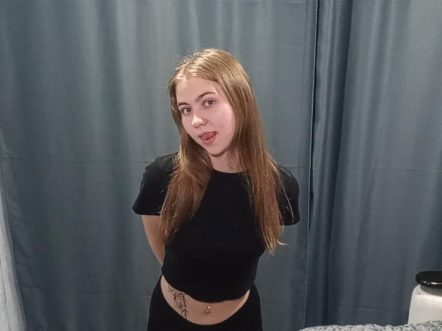 MargretWarriner has long blonde hair, black t-shirt, black pants, and a belly button piercing, standing with arms behind her back, looking at the camera with a slight pout.