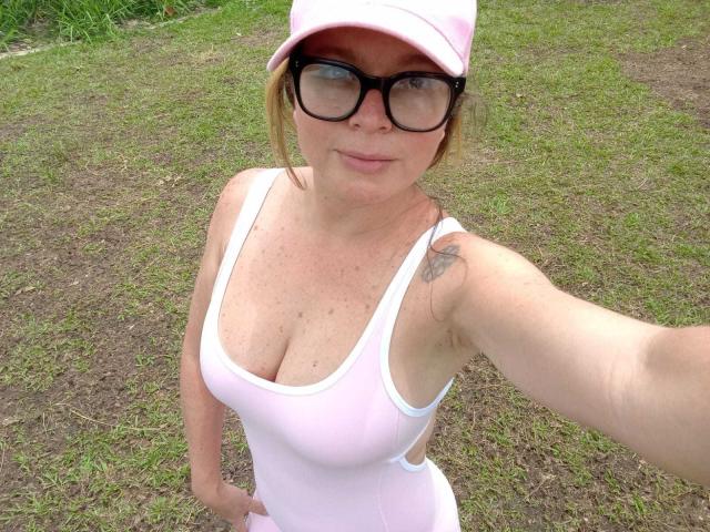 PamelaColin con gli occhiali, un berretto rosa e una canottiera bianca che mette in mostra un seno prosperoso, sfondo esterno con erba, tatuaggio sul braccio destro, che tiene in mano il telefono per un selfie..