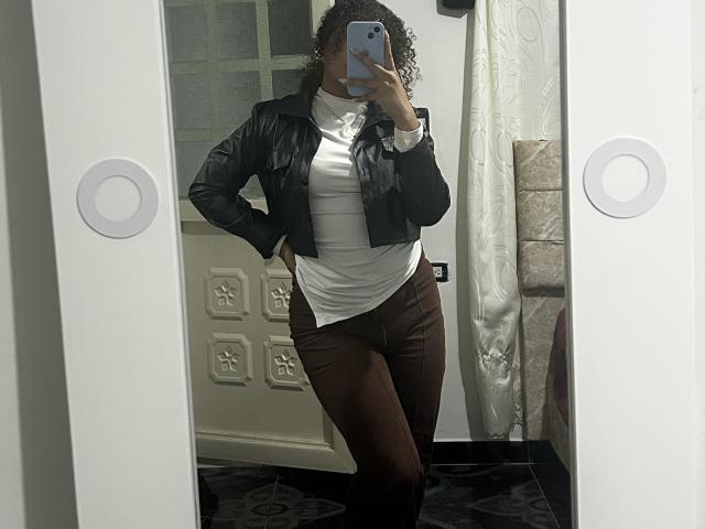Sofia, eine Frau mit lockigen Haaren, trägt eine schwarze Lederjacke, ein weißes Hemd und braune Hosen. Sie steht vor einem Spiegel und macht ein Selfie, wobei eine Hand an der Hüfte liegt und das Telefon vor ihr gehalten wird. Im Hintergrund sind eine weiße Tür und Wanddekoration zu sehen..