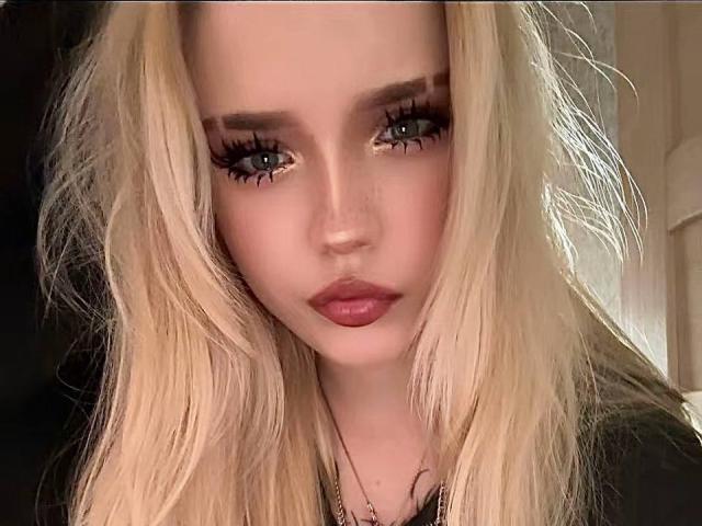SaturninaFischbein has blonde hair, blue eyes, and pink lipstick. She's staring directly at the camera with long, thick eyelashes. Her face is centered in the shot.