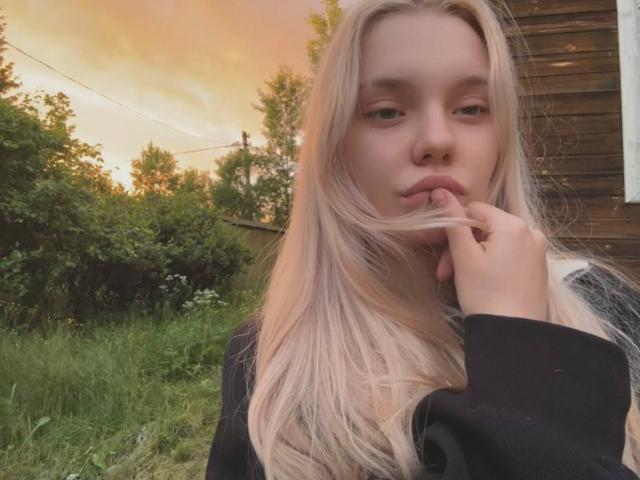 Saturnina Fischbein, met lang blond haar, lichte huid en volle lippen, die aan haar vinger knabbelt, draagt een zwarte top en staat in een grasveld bij zonsondergang..