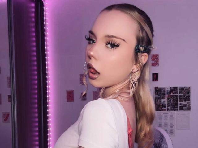 Saturnina Fischbein, met blond haar, lange wimpers en volle lippen, draagt een witte top, kijkt over haar schouder, met een paarse LED-strip aan de linkerkant, oorbellen, in een zwak verlichte kamer..
