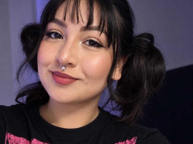 IsabellArt ha due trecce scure, un piercing al naso e indossa una maglietta nera con dettagli rosa. Sorride, con il rossetto rosa e le lunghe ciglia. La sua pelle è chiara e liscia..