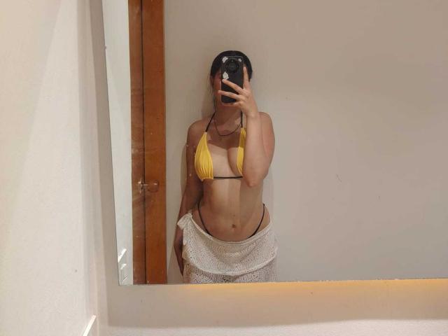 ViolethaMoretti taking a mirror selfie, wearing a yellow bikini top that barely covers her small tits, and a white mesh skirt, with her hand covering her phone.