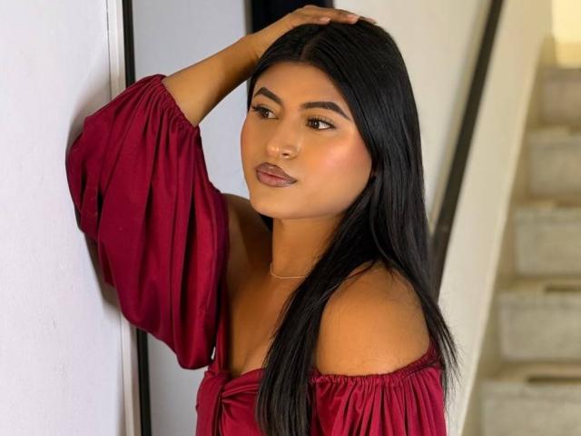 MichelleNarvaez with long black hair, wearing a red off-shoulder dress, hand in hair, looking at camera, brown eyes, subtle makeup, soft smile, standing against white wall.