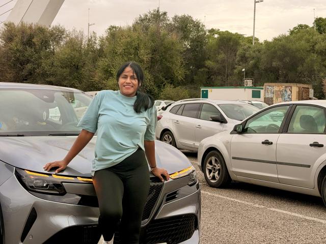 IvannaDuran69 leans against a silver car in a parking lot, wearing a light blue shirt and black pants, smiling with dark hair and medium brown skin.