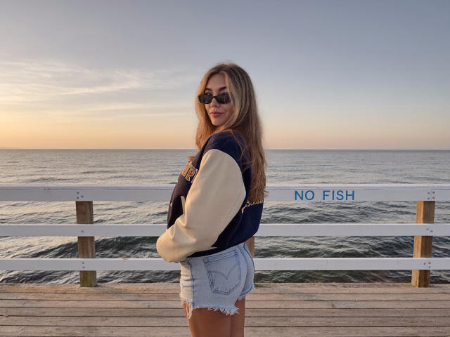 Sofie White mit langen, blonden Haaren, die dunkle Sonnenbrillen trägt, eine dunkelblaue und beigefarbene Sportjacke und zerrissene, blaue Jeansshorts, steht auf einem hölzernen Steg bei Sonnenuntergang..