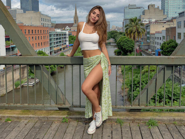 Sofie White, con lunghi capelli castani, canotta bianca, gonna verde a fiori con spacca alta che mostra il sedere, scarpe da ginnastica bianche, appoggiata alla ringhiera di un ponte, con sullo sfondo il panorama urbano..