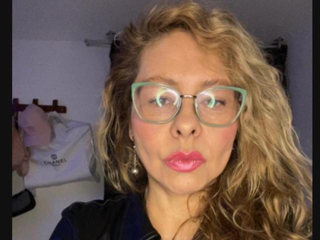 MiaAmoretti, con i capelli biondi ricci, gli occhiali verdi e il rossetto rosa, che fa una smorfia, indossa una maglietta nera e porta degli orecchini pendenti, in un selfie..