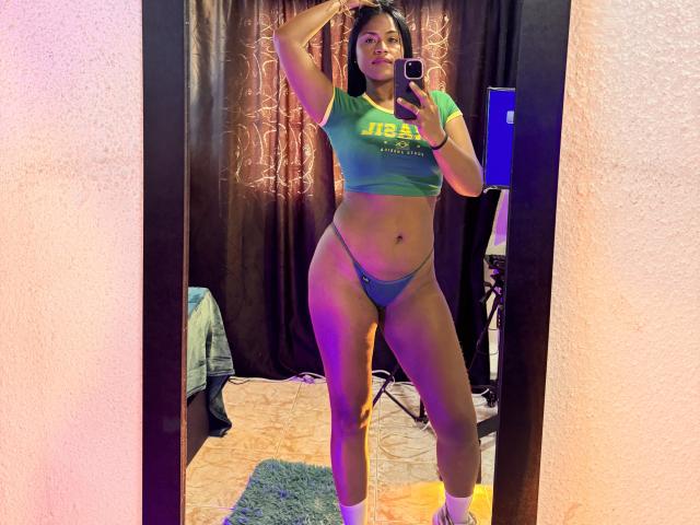 IvannaDuran69 with curvy, thick thighs, small tits in green crop top, blue thong, standing in front of mirror, holding phone, pink wall, dark curtains, blue carpet.