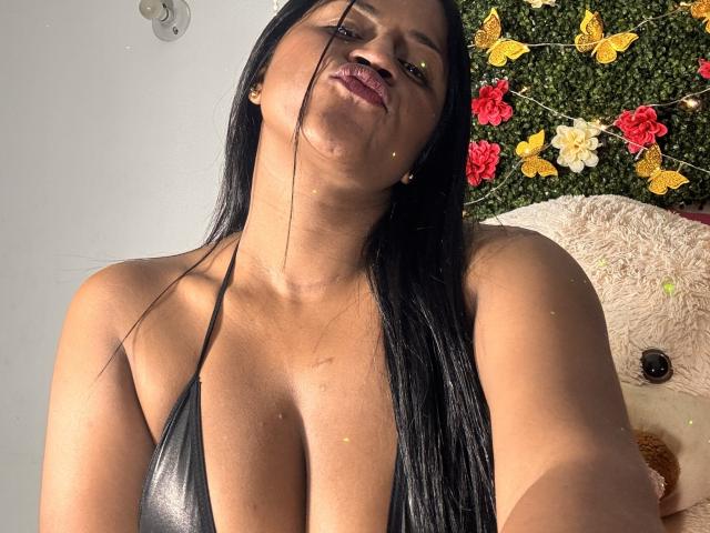 IvannaDuran69 with long black hair, medium brown skin, wearing a black halter top, showing her large tits, slightly open mouth, and a small yellow dot on her cheek.
