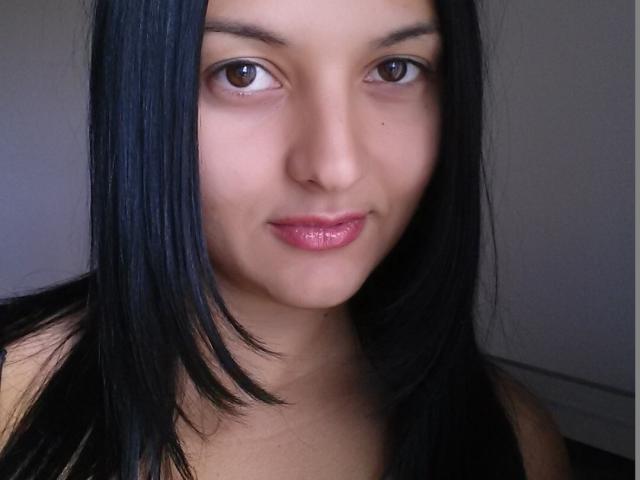 Desireey with shiny black hair, brown eyes, and pink glossy lips, staring into the camera with a slight smile, showing her smooth, tan-skinned face and bare shoulders.