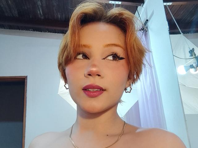 Chloee Williams a les cheveux courts et orange, les yeux marron et du rouge à lèvres rose. Elle porte de petites boucles d'oreilles et un collier, et regarde légèrement sur le côté avec un sourire discret..