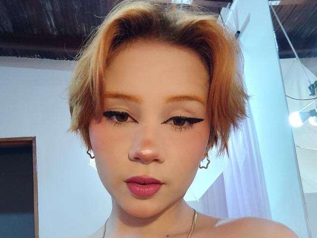 ChloeeWilliams with short, dirty-blonde hair, cat-eye eyeliner, pink lips, and small hoop earrings, looking down at the camera with a neutral expression. She's wearing a silver necklace.
