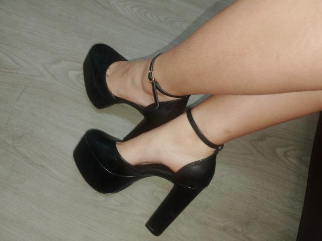 Les jambes de Chloee Williams sont chaussées de chaussures noires à talons hauts et compensées, avec des brides aux chevilles, et elle se tient sur un sol en bois clair. Sa peau nue, lisse et bronzée contraste avec le cuir noir brillant..