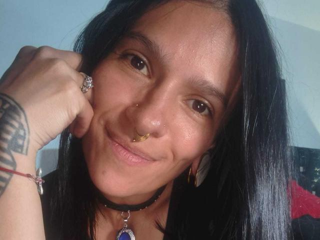 AmelieBlake a un petit anneau au nez, un collier noir avec un pendentif violet, des cheveux noirs, un tatouage sur l'avant-bras gauche, et elle sourit avec ses yeux marrons. Elle porte une bague et des boucles d'oreilles..
