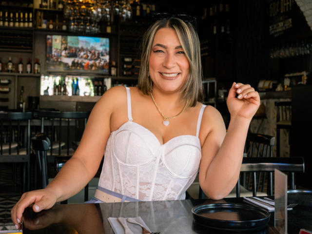 EmaFoxy met een gemiddelde borstmaat, draagt een witte kant bustier, glimlachend, met een drankje in haar hand, en heeft zwarte zonnebrillen op haar hoofd, in een bar met een televisie op de achtergrond..