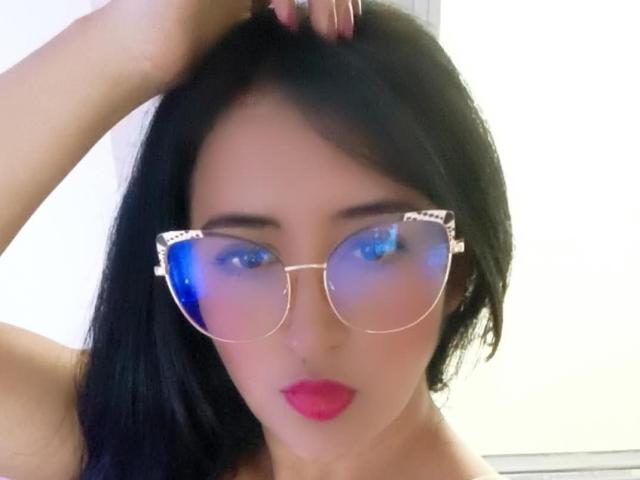 NicollJeans has black hair, blue reflective glasses, pink lipstick, and a hand in her hair. She's looking at the camera with a serious expression.