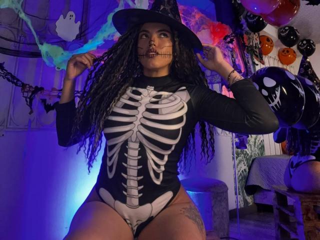 LiliRose69 in a skeleton-themed bodysuit with black sleeves, wearing a witch hat, holding her hat, with long curly hair, purple-blue lighting, and Halloween balloons in the background.