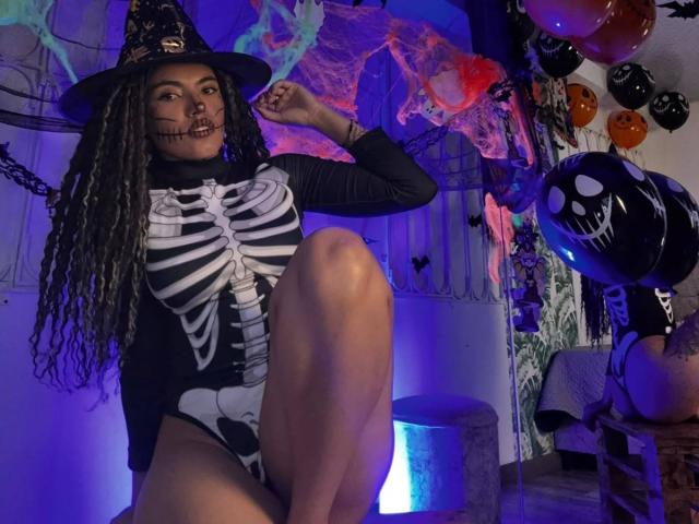 LiliRose69 with long curly hair, wearing a black witch hat, skeleton bodysuit, and black mask, sitting with leg raised, surrounded by Halloween balloons and decorations.