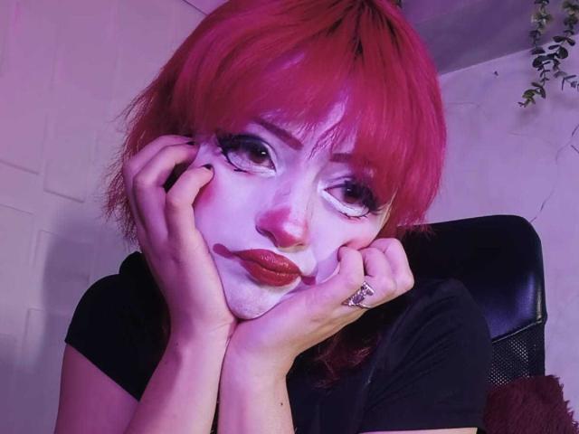 ViolettClay con i capelli rosa, il viso dipinto di bianco e il rossetto rosso, le mani sulle guance, una maglietta nera, un anello, illuminazione viola-rosa, che guarda verso l'alto, capelli leggermente disordinati, espressione dolce, interni..