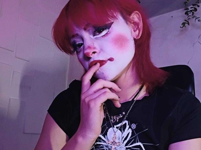 ViolettClay with red hair, pale makeup, pink blush, black shirt, finger to mouth, looking seductive, purple light, necklace with spider design.