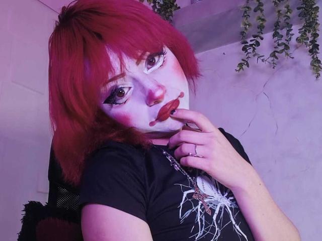 ViolettClay, avec ses cheveux roux, son maquillage blanc sur le visage et son rouge à lèvres rouge, touchant son menton avec un doigt, portant une chemise noire avec un motif d'araignée, dans une pièce éclairée en violet..