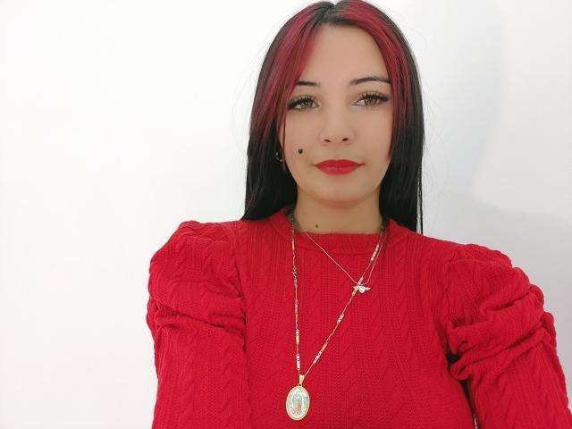 Xhimena with red and black hair, red lipstick, red sweater, and a necklace with a round pendant, smiling with a small beauty mark on her cheek.