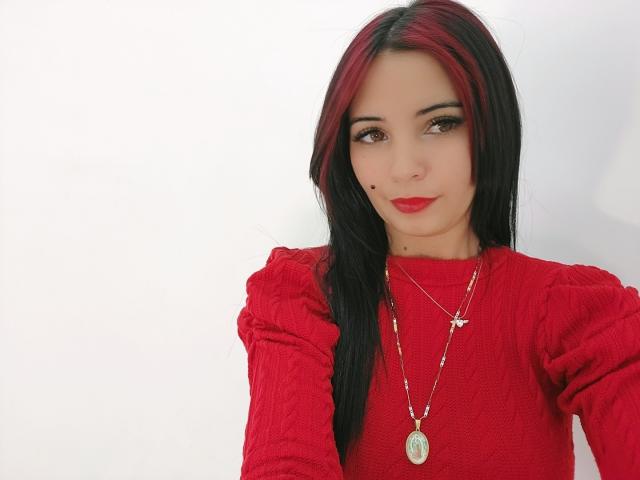Xhimena, avec ses cheveux noirs et rouges, son pull rouge et ses deux colliers, montre une adorable petite tache de rousseur sur sa joue, et sourit avec du rouge à lèvres. Sa poitrine n'est pas visible, seulement le haut de son corps..