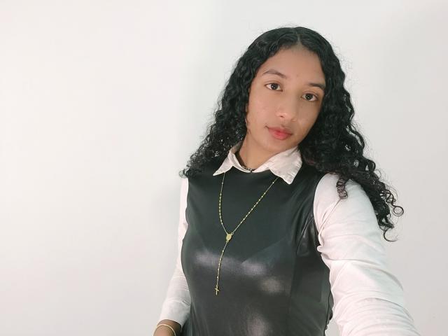 Alhara, met krullend zwart haar, draagt een zwart leren vest over een wit shirt, een gouden ketting en een gouden armband, en kijkt met een lichte glimlach naar de camera..