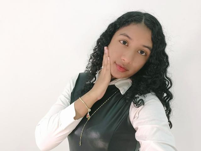 Alhara, avec ses cheveux noirs bouclés, portant une chemise blanche et un gilet en cuir noir, touche sa joue avec sa main gauche, un collier avec une croix dorée, un sourire doux, une peau claire..