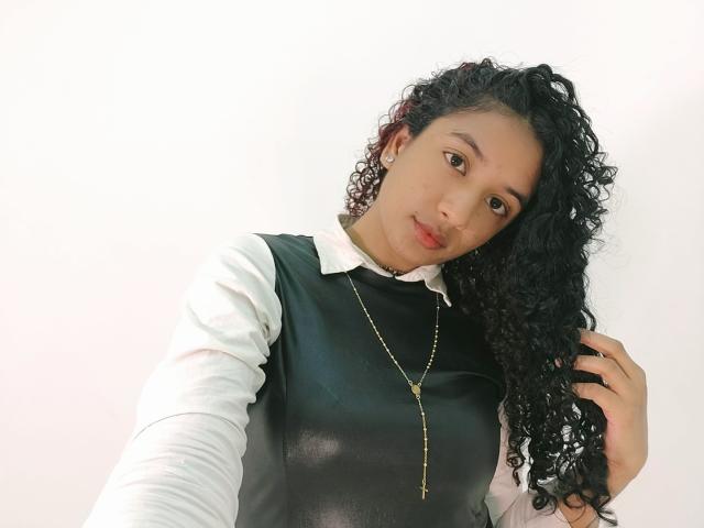 Alhara, com cabelos pretos e cacheados, vestindo uma camisa preta e branca, um colar com uma cruz dourada e brincos, segurando o cabelo, olhando para a câmera com um leve sorriso..